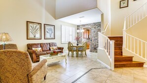 A spacious living room with a brown leather sofa, patterned armchair, and a dining area with a table and chairs. A staircase with white railings leads to an upper level. - Spacious 5-bedroom house 3 min from Cardinals Stadium and West Gate shopping (Phoenix)
