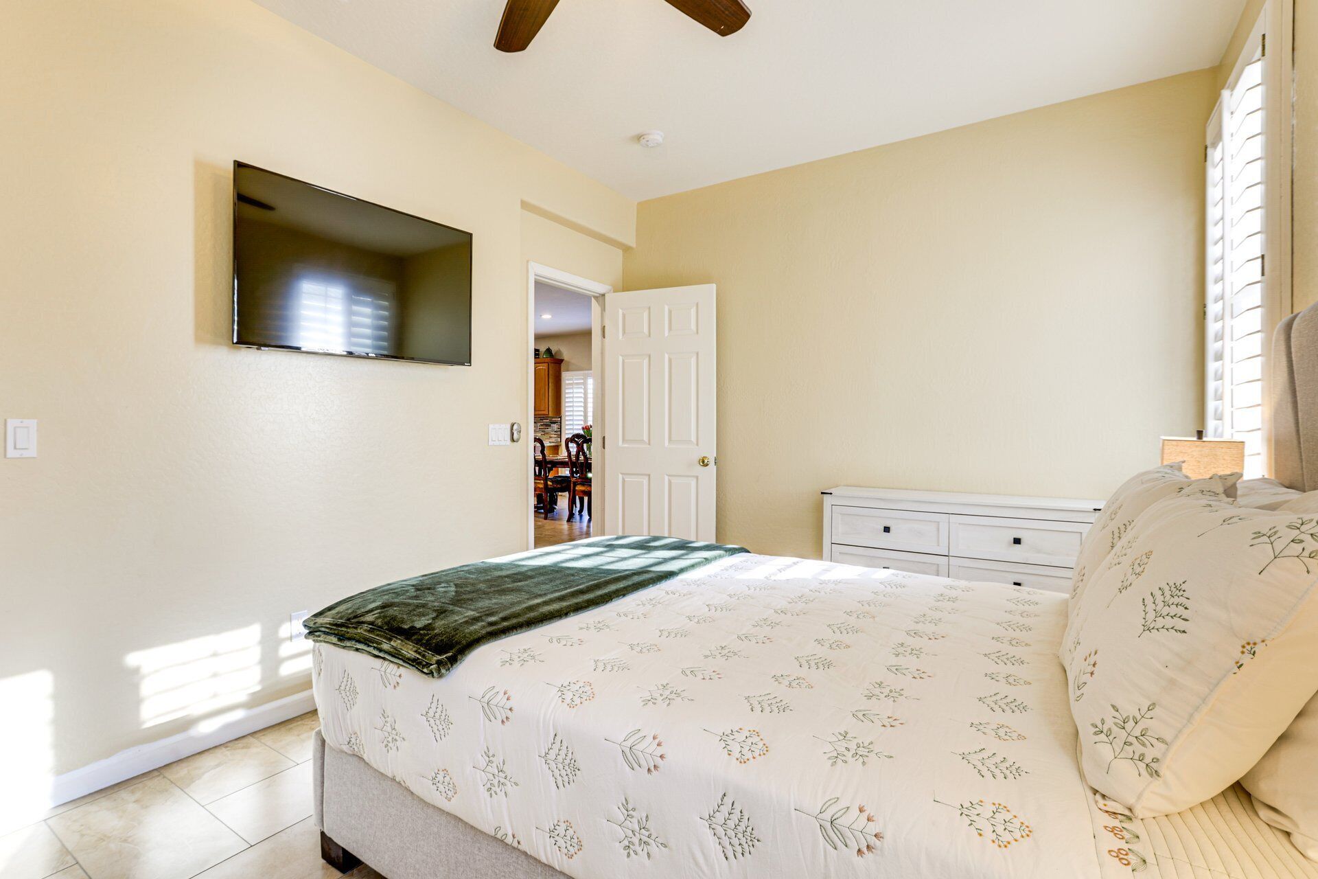 Un dormitorio con cama, televisor montado en la pared, puerta que da al baño y ventana con persianas.