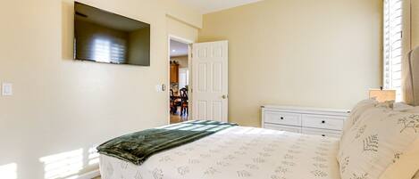 Un dormitorio con cama, televisor montado en la pared, puerta que da al baño y ventana con persianas.