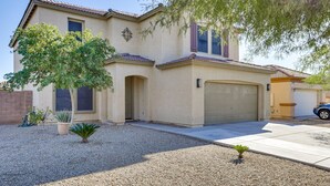 A single-story house with a beige stucco exterior, a tiled roof, and a garage. There are two windows, one with shutters, and a small tree in the front yard. - Spacious 5-bedroom house 3 min from Cardinals Stadium and West Gate shopping (Phoenix)