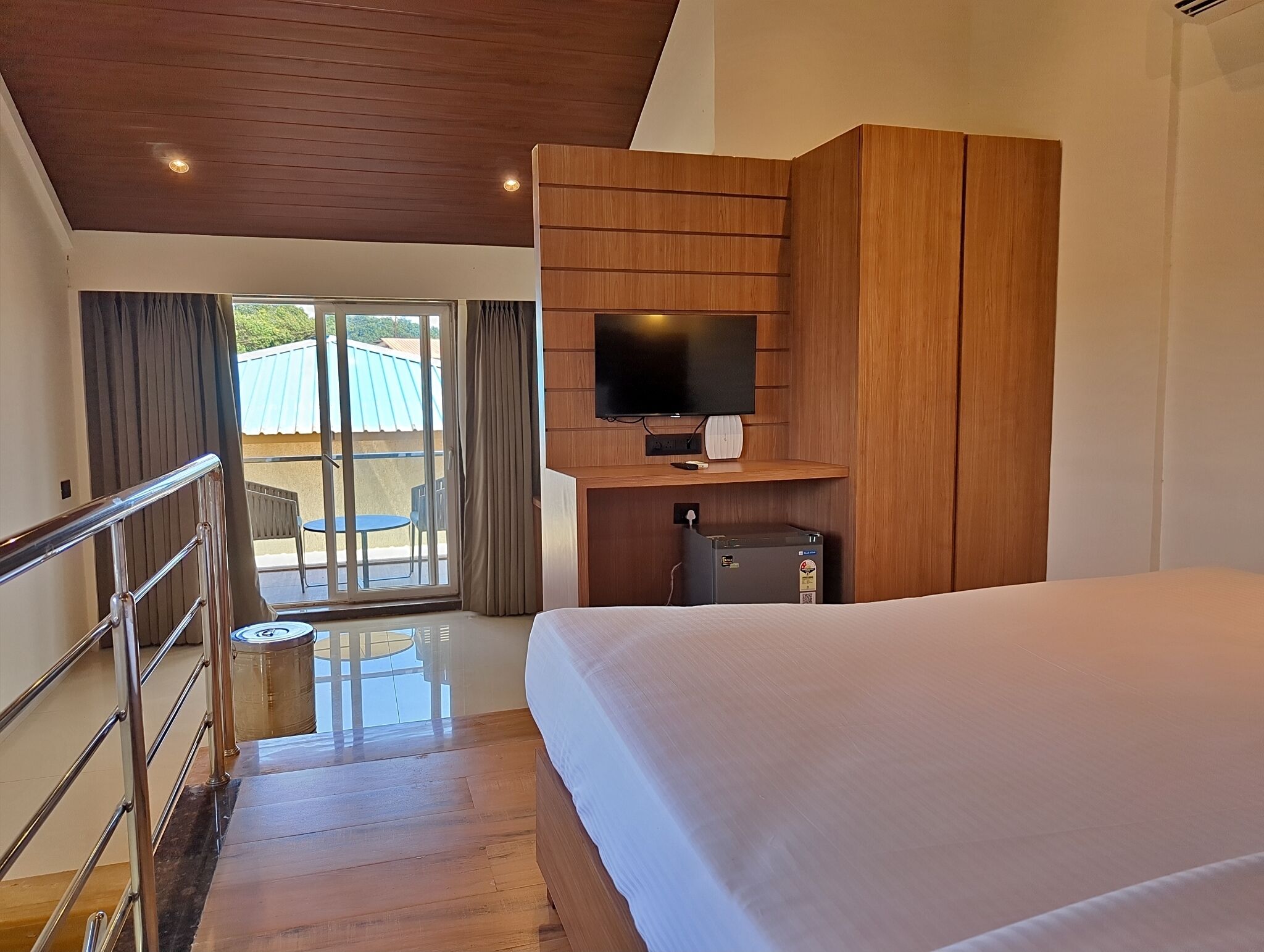 A hotel room with a large bed, wooden ceiling, a flat-screen TV mounted on a wooden panel, and a sliding glass door leading to an outdoor area.