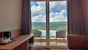 Una habitación con un escritorio de madera, una silla y una mesa con una bandeja de vasos y una tetera. Un ventanal ofrece vistas a un balcón con sillas y una mesa, con vistas a un paisaje verde y un puente.