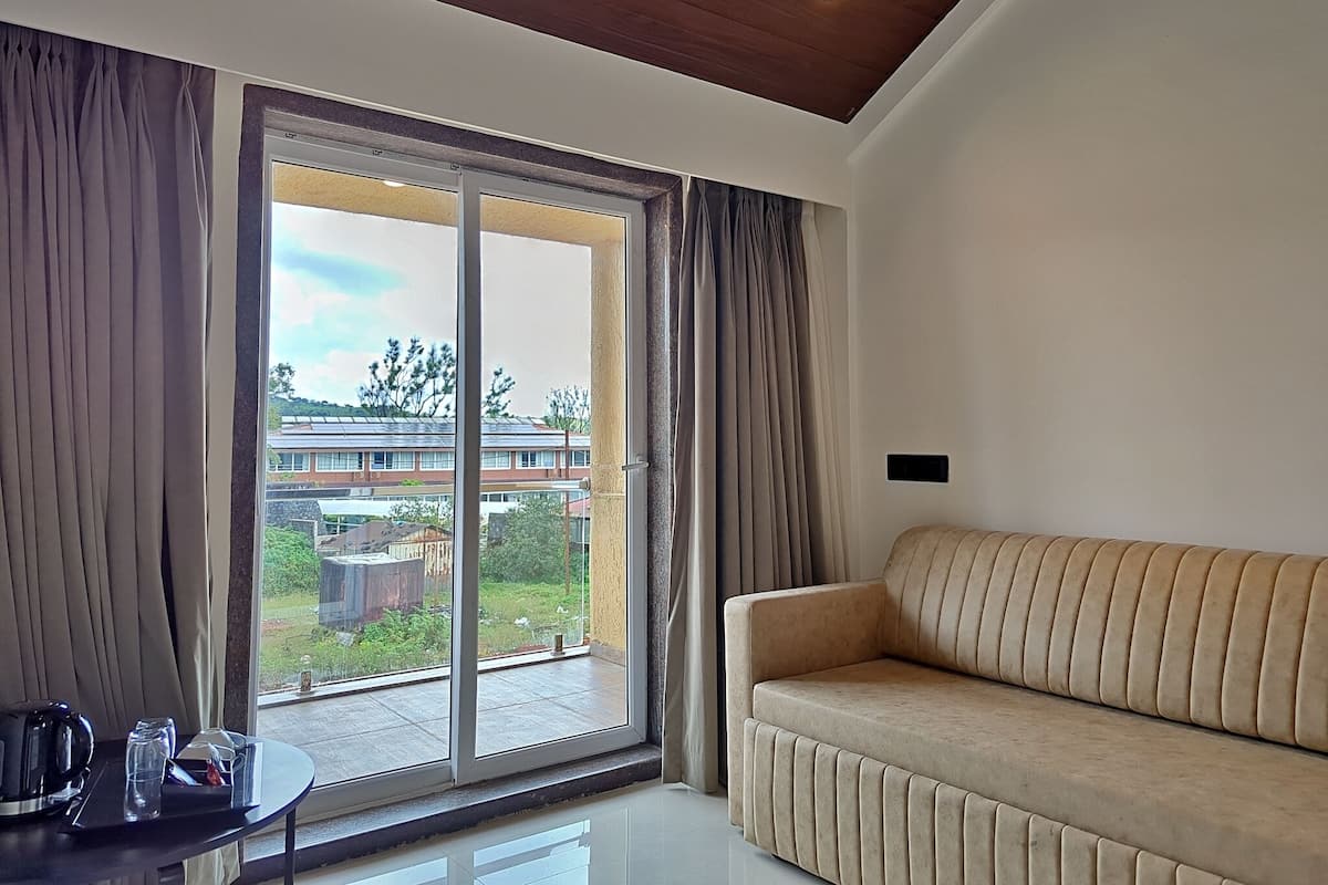 Una sala moderna con un sofá, una mesa con hervidor y vasos, y un ventanal corredizo que da a un balcón con vistas a edificios y vegetación.