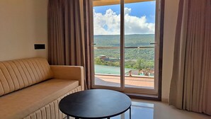 Una habitación con sofá y una mesita, un ventanal con cortinas y vistas a un paisaje verde.