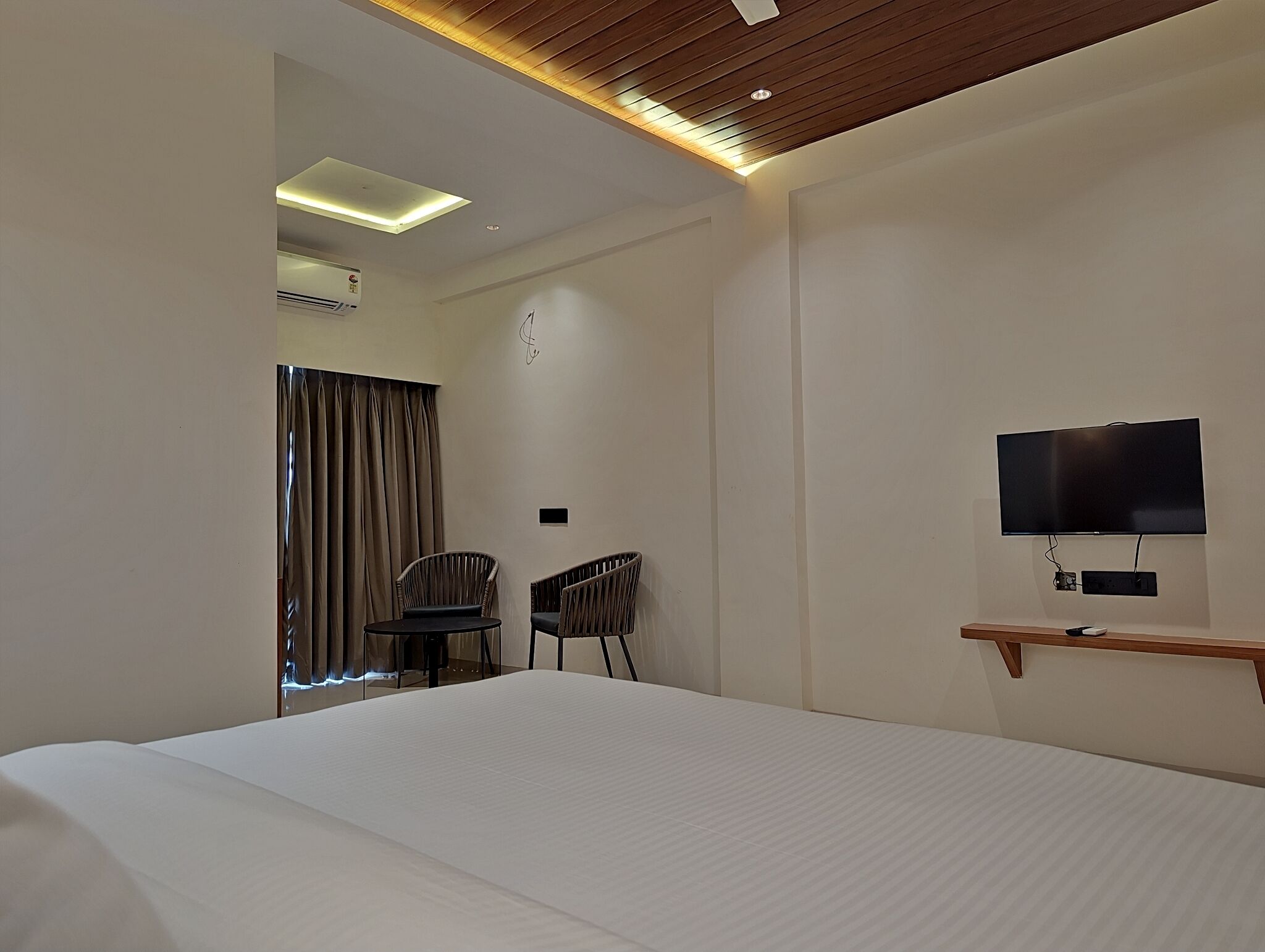 A hotel room with a large bed, a TV mounted on the wall, a desk with two chairs, and a window with curtains.