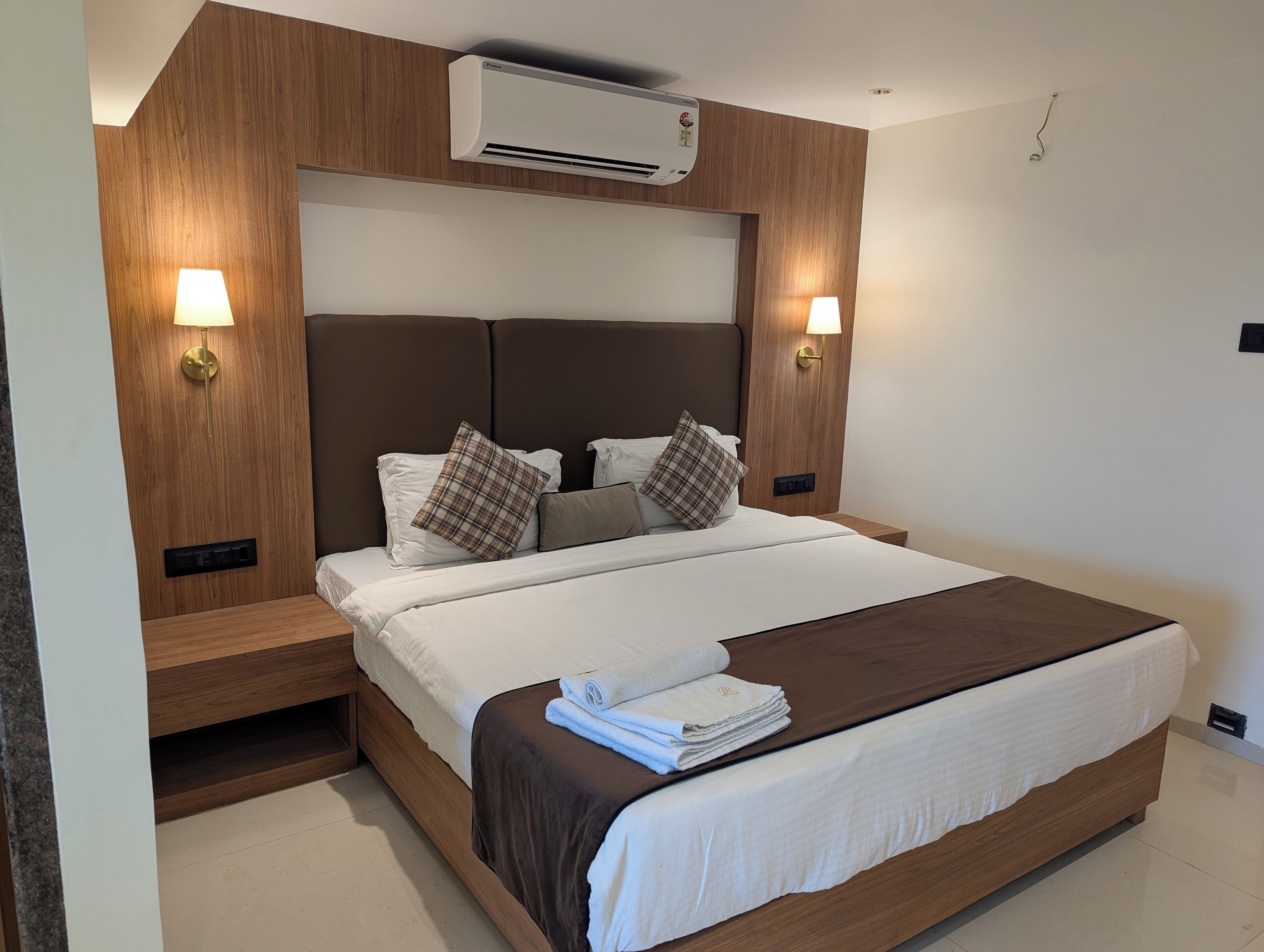 A modern bedroom with a large bed, wooden headboard, two bedside lamps, and an air conditioning unit.