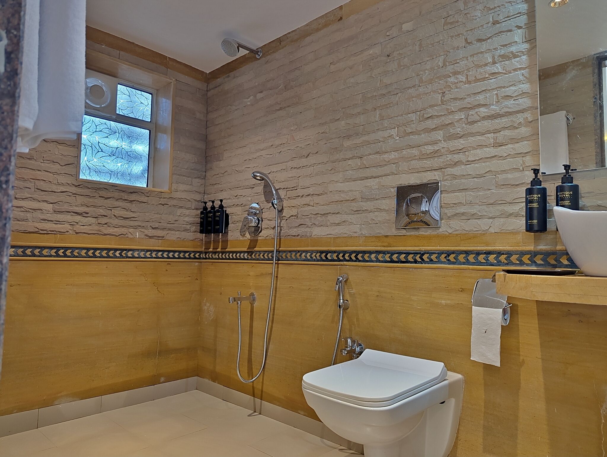 Un baño con ducha, inodoro y lavabo. Se ve una ventana con vistas a árboles, y hay artículos de tocador visibles en la pared.