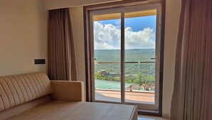 Habitación de hotel con una cama grande, un sofá y un ventanal corredero que da a un balcón con vistas al paisaje.