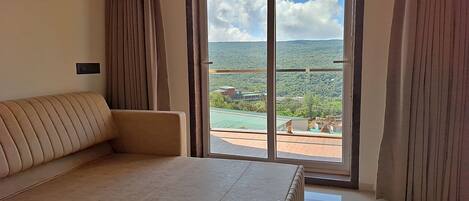 A hotel room with a large bed, a sofa, and a sliding glass door leading to a balcony with a view of the landscape.