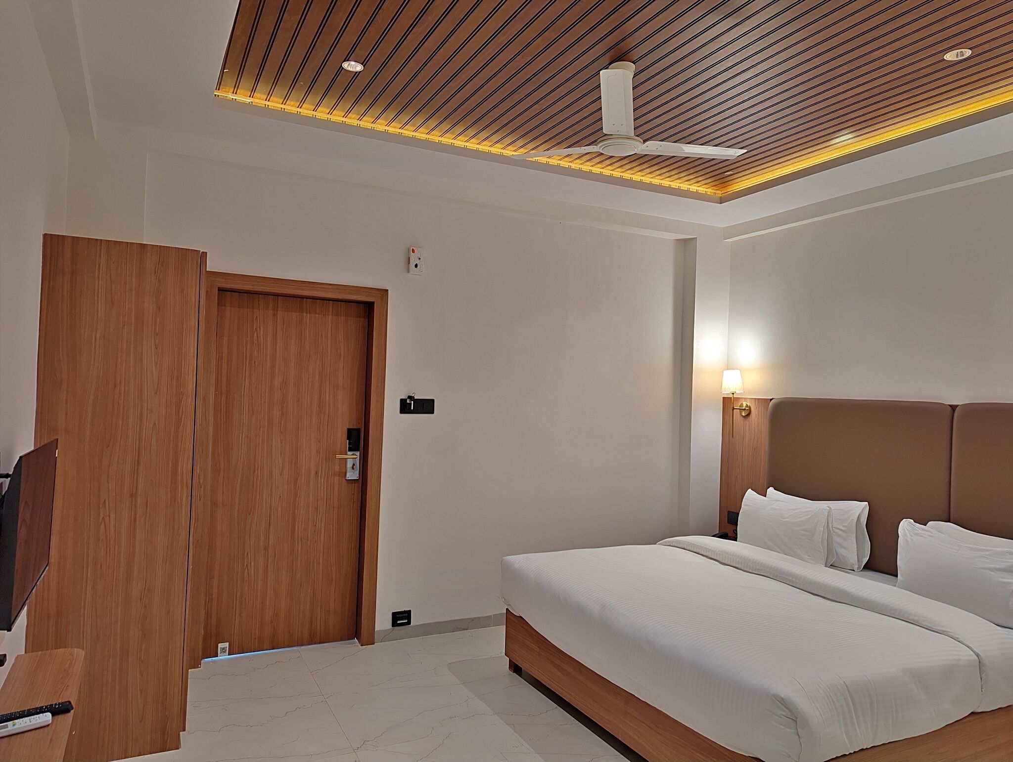 A modern hotel room with a wooden door, a bed with white linens, a ceiling fan, and recessed lighting.