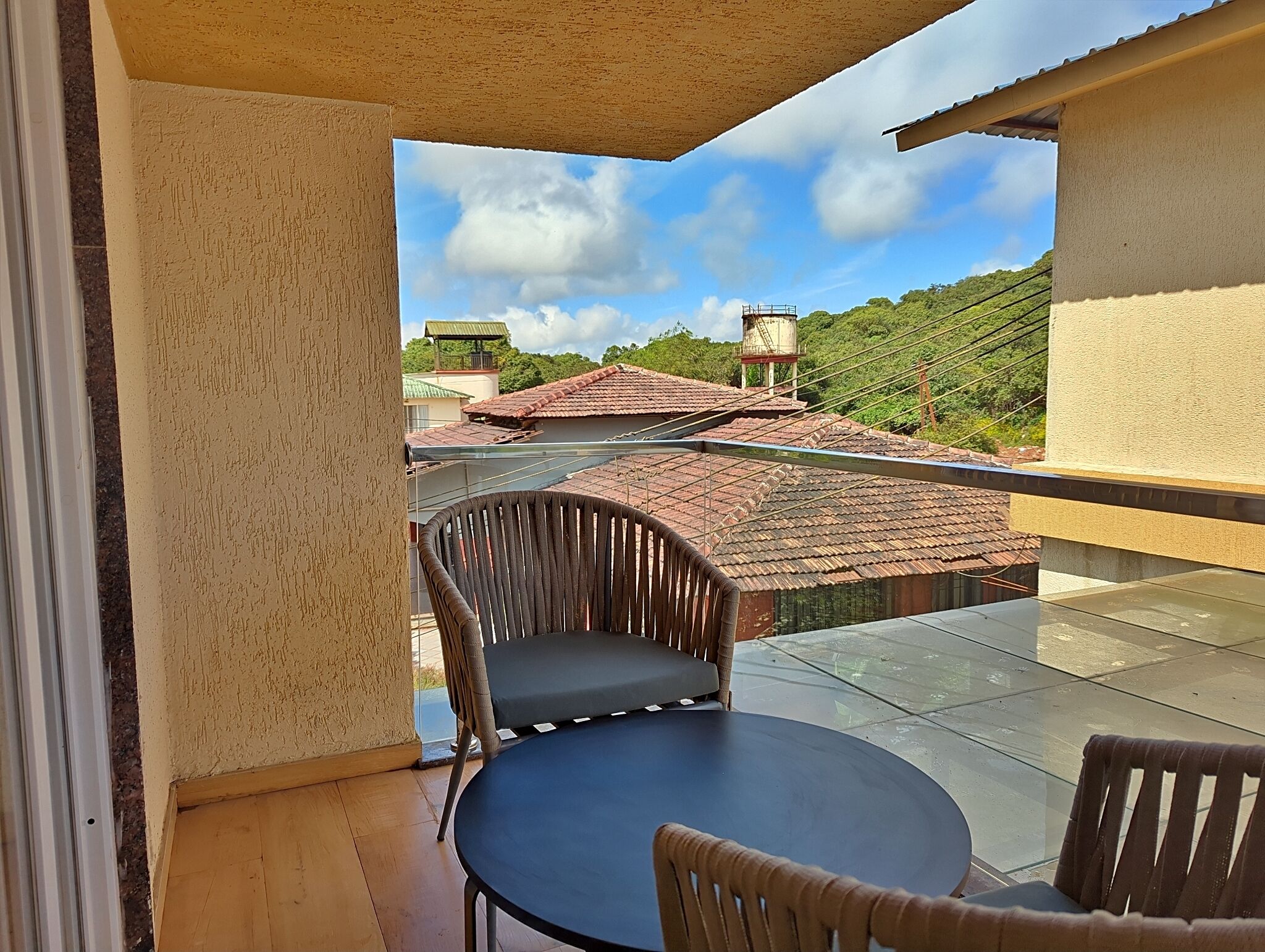 A balcony with a round table and two chairs, overlooking a residential area with houses and a hill.