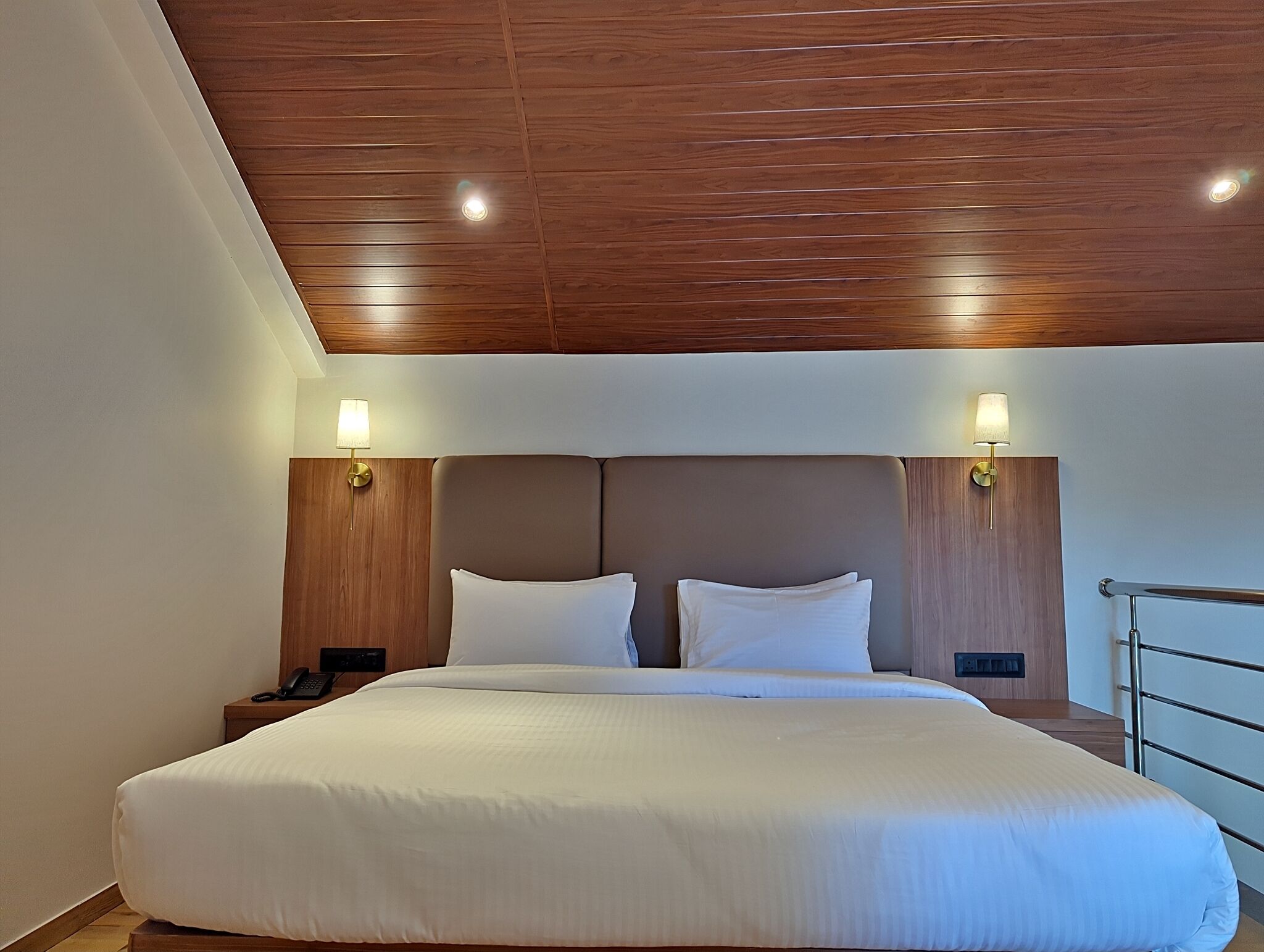 A hotel room with a large bed, wooden ceiling, two bedside lamps, and a nightstand with a phone.