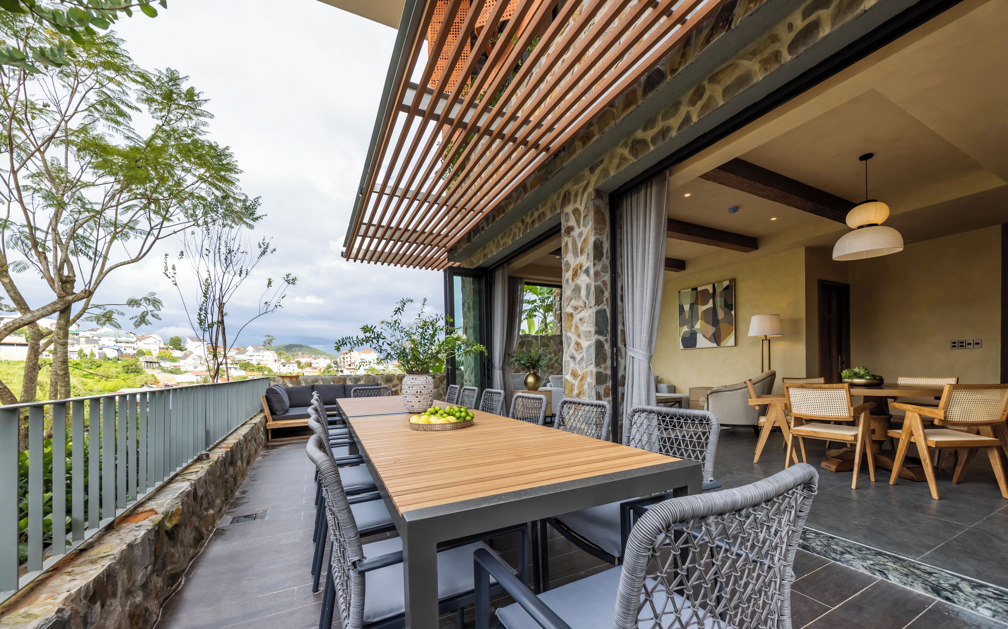 A dining area with a long wooden table and wicker chairs, a stone wall, and a view of the cityscape.