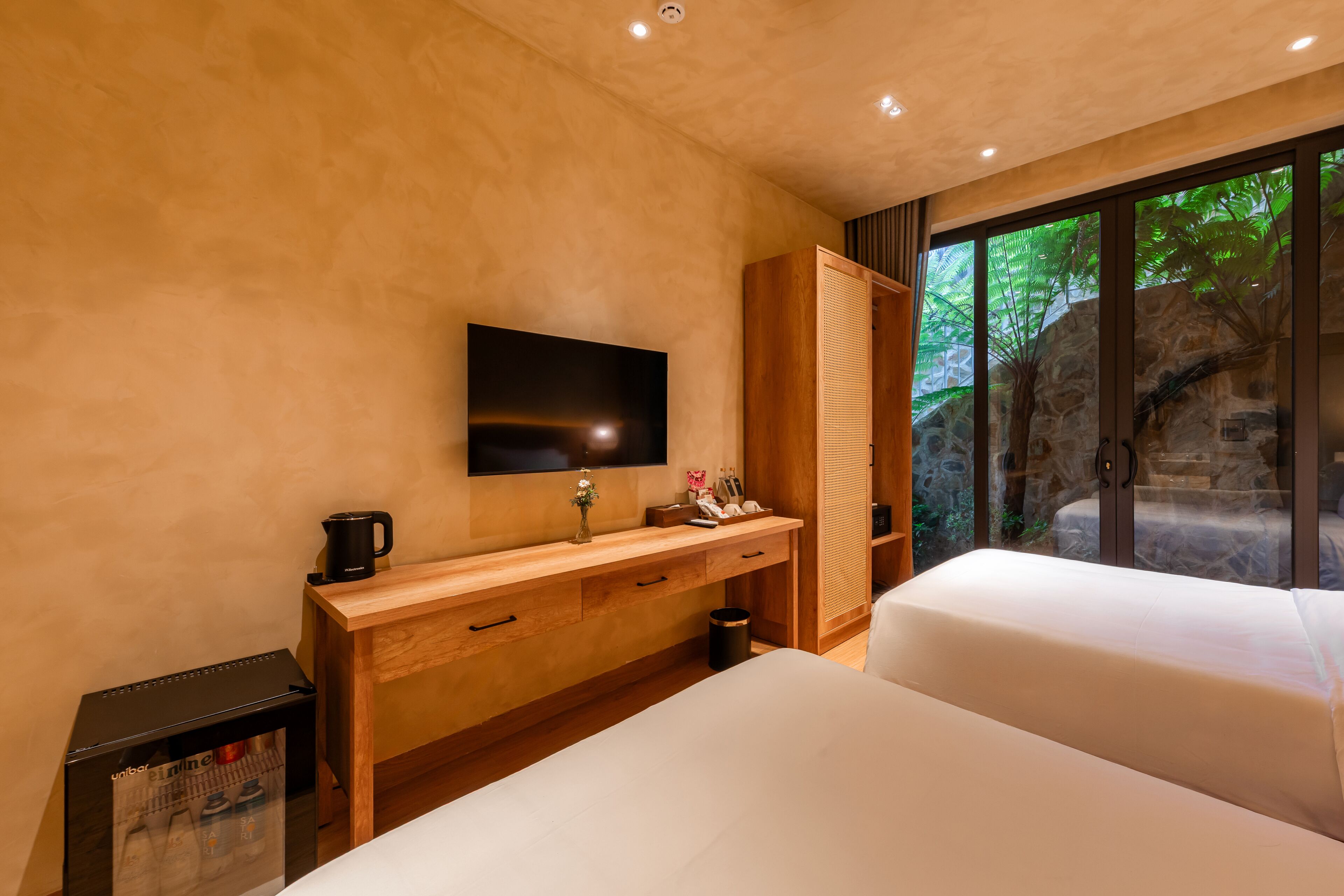 A modern hotel room with a large bed, a flat-screen TV mounted on the wall, a wooden console table with drawers, a small trash bin, and a view of a stone wall and greenery through a glass door.