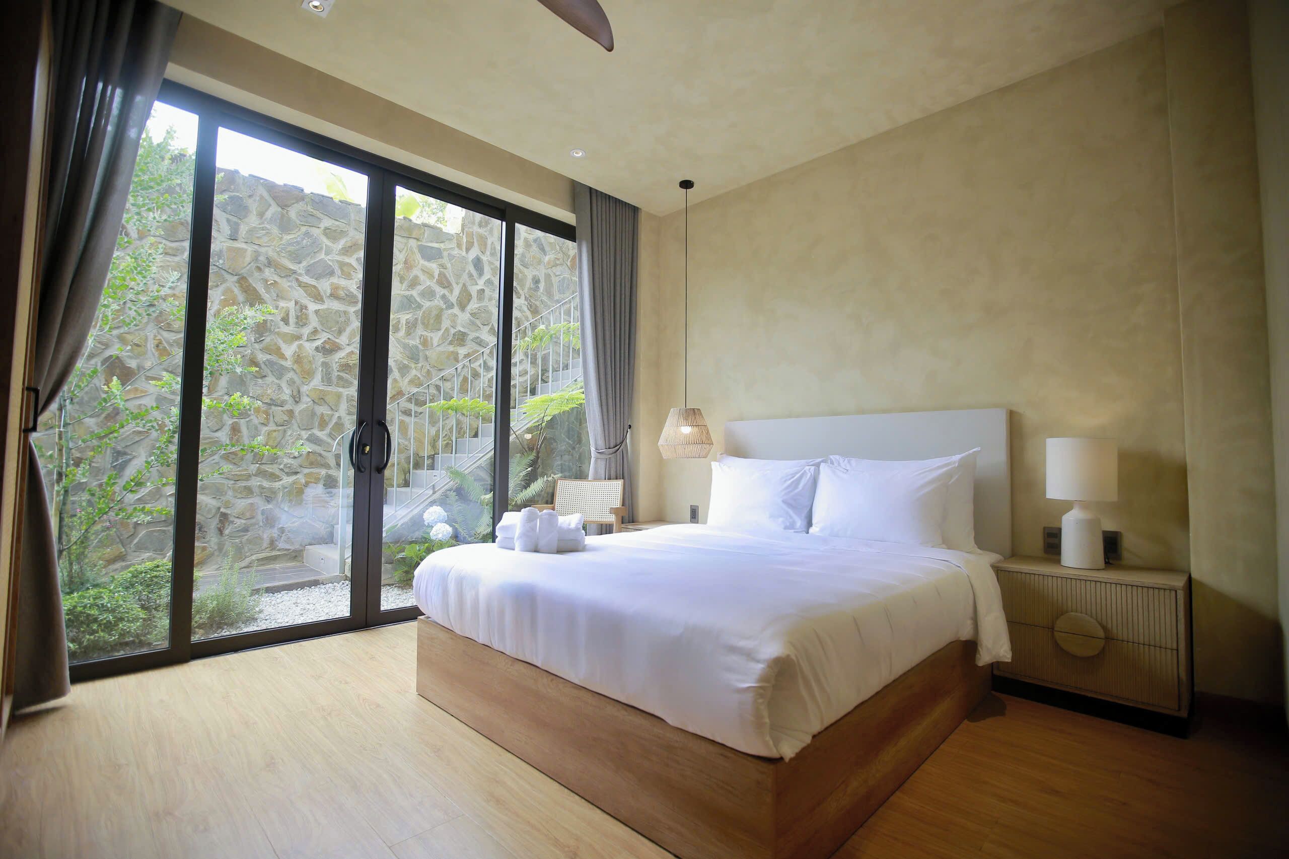 A bedroom with a large bed, a nightstand with a lamp, and a view of a stone wall and greenery outside.