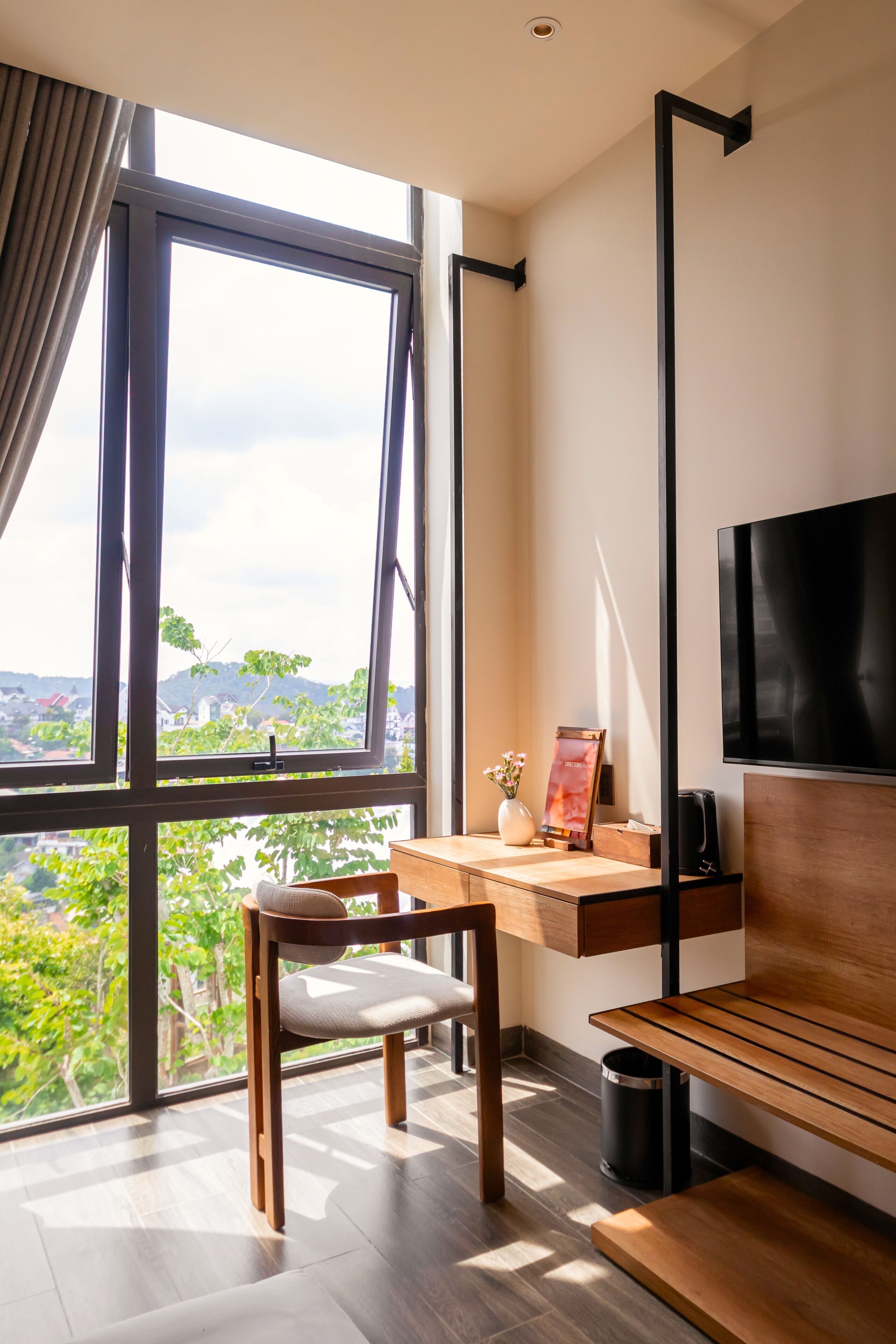 Habitación moderna con un amplio ventanal, un escritorio, una silla, un televisor y un pequeño cubo de basura.