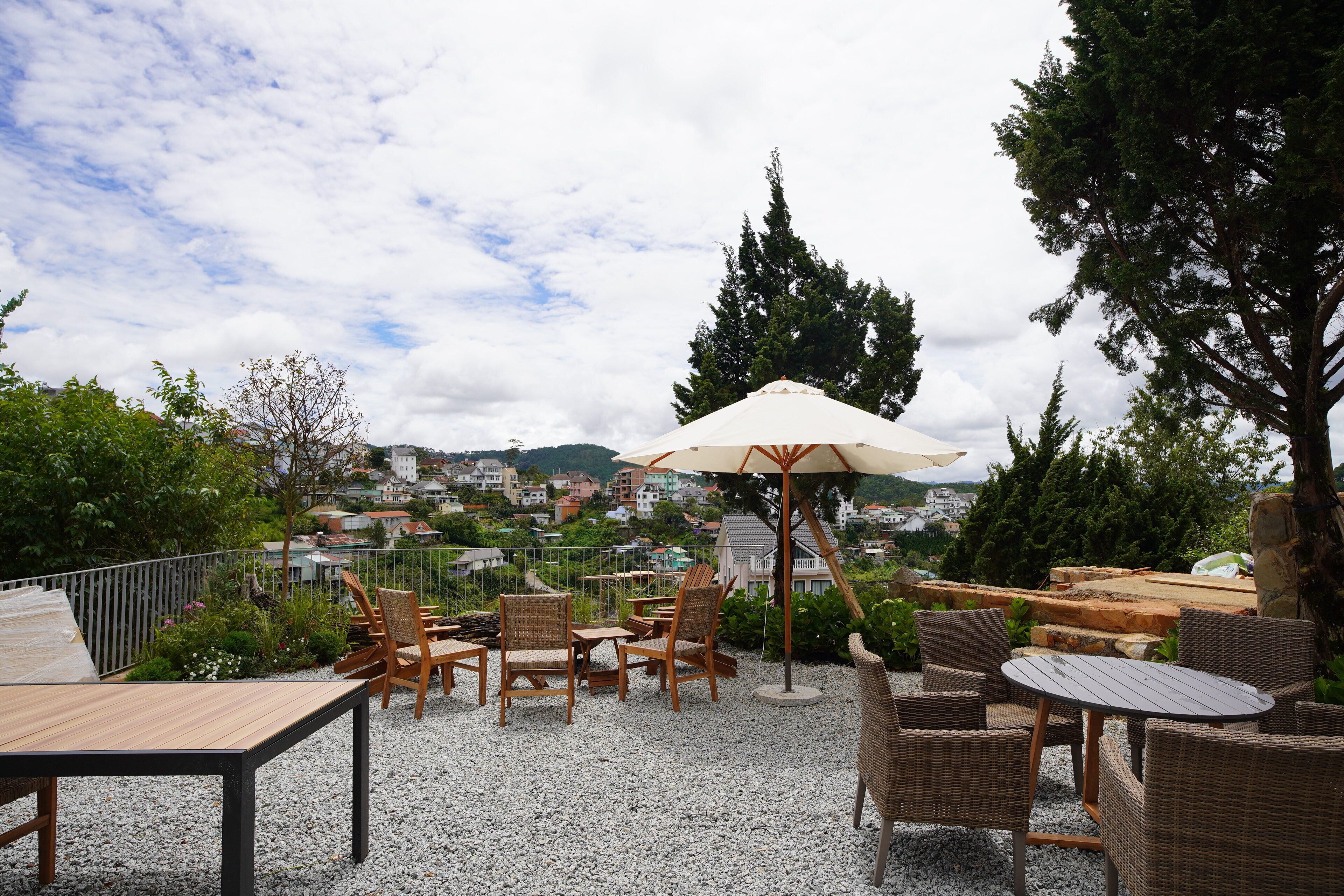 An outdoor seating area with wicker furniture and a large umbrella, overlooking a town with houses and trees.