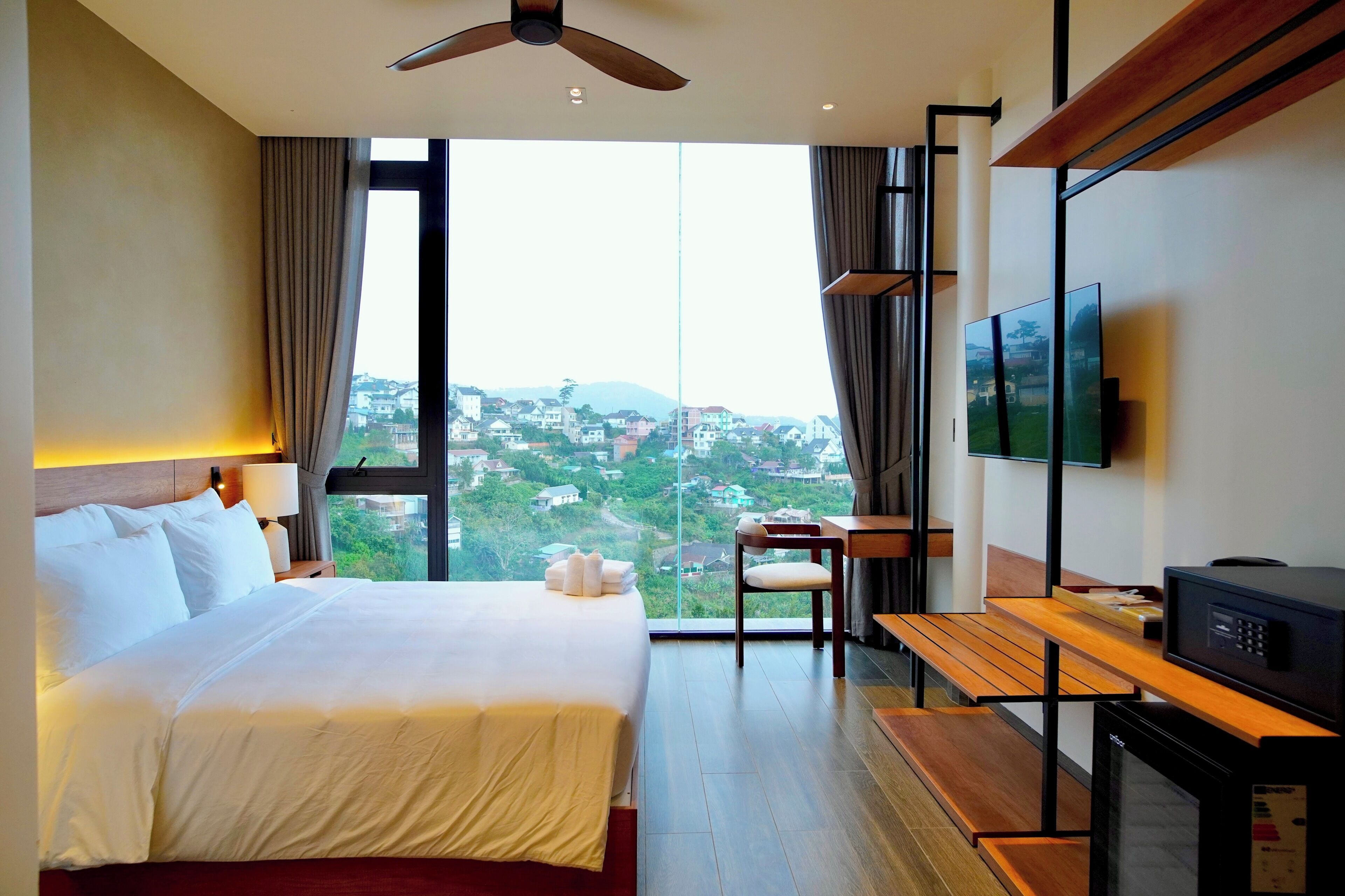 A hotel room with a bed, a desk, a chair, a television, and a view of a cityscape.