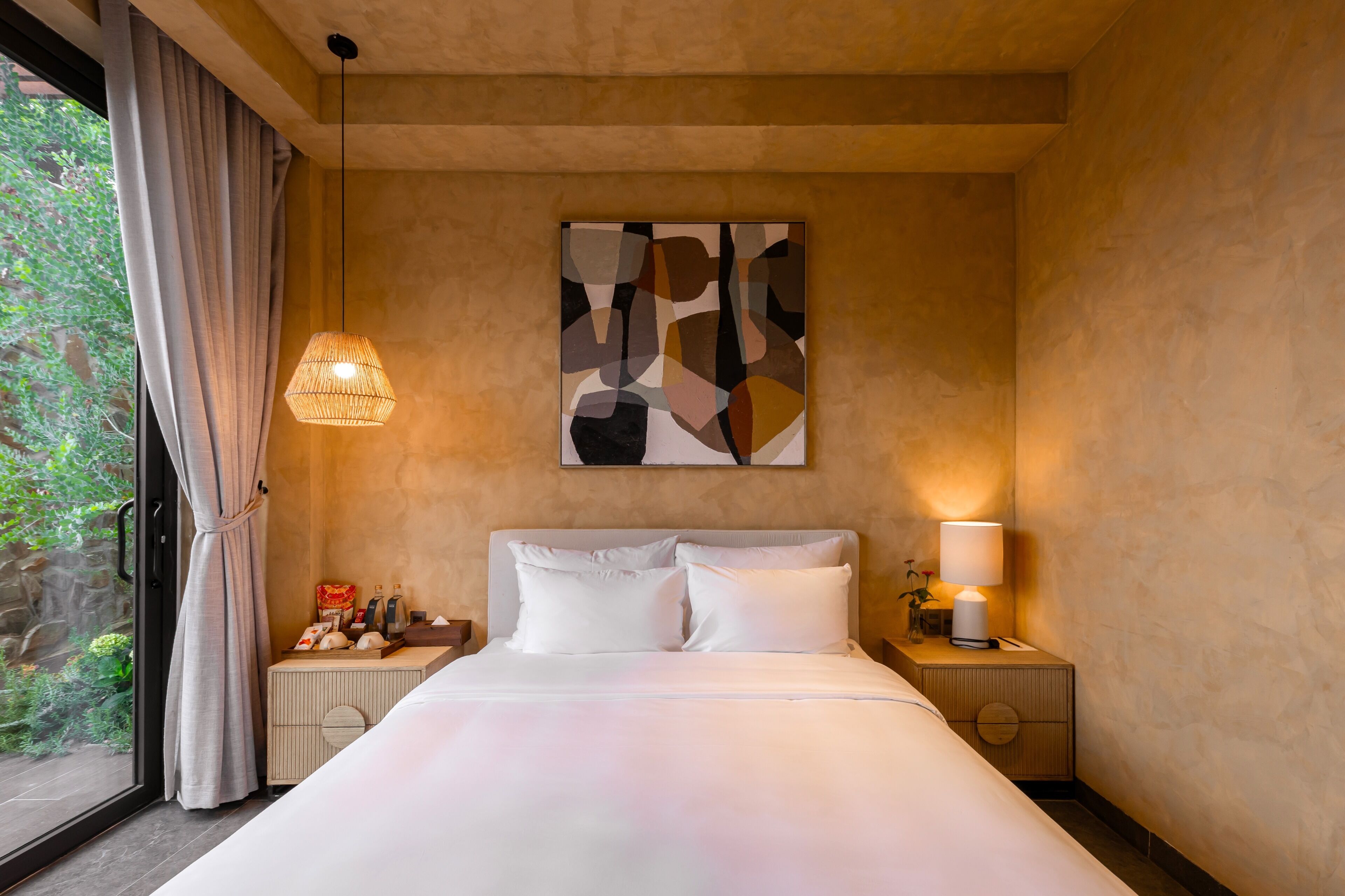 Een hotelkamer met een bed, nachtkastjes, een lamp, een schilderij en een raam met gordijnen.