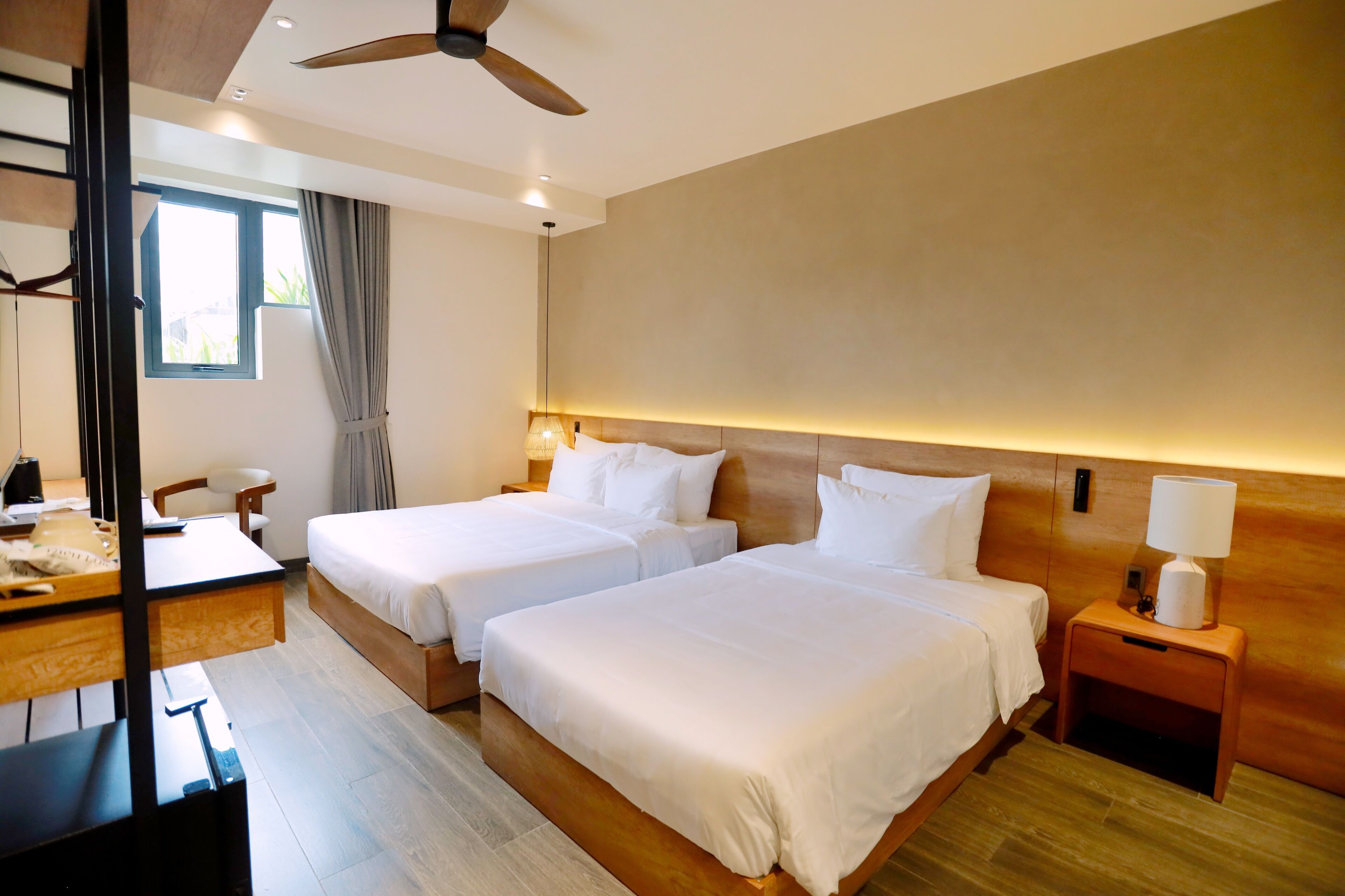 Habitación de hotel con dos camas, un escritorio, una silla, un ventilador de techo y una ventana con cortinas.
