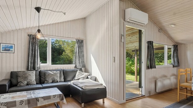 A modern living room with a sectional sofa, a coffee table, and a dining area with a chair. There is a large window with curtains, an air conditioning unit, and a door leading outside.