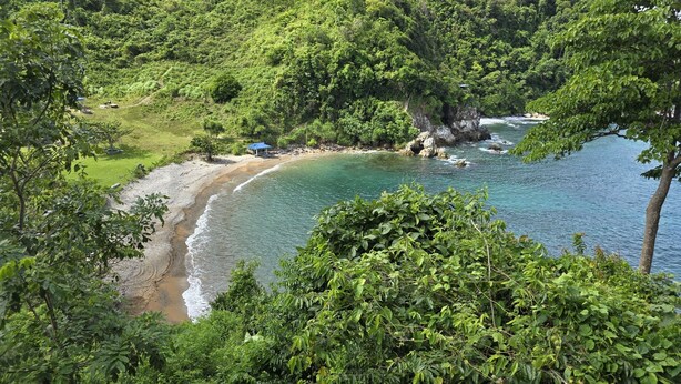 Un paysage côtier avec une plage, une petite construction dotée d’un toit bleu et une végétation luxuriante.