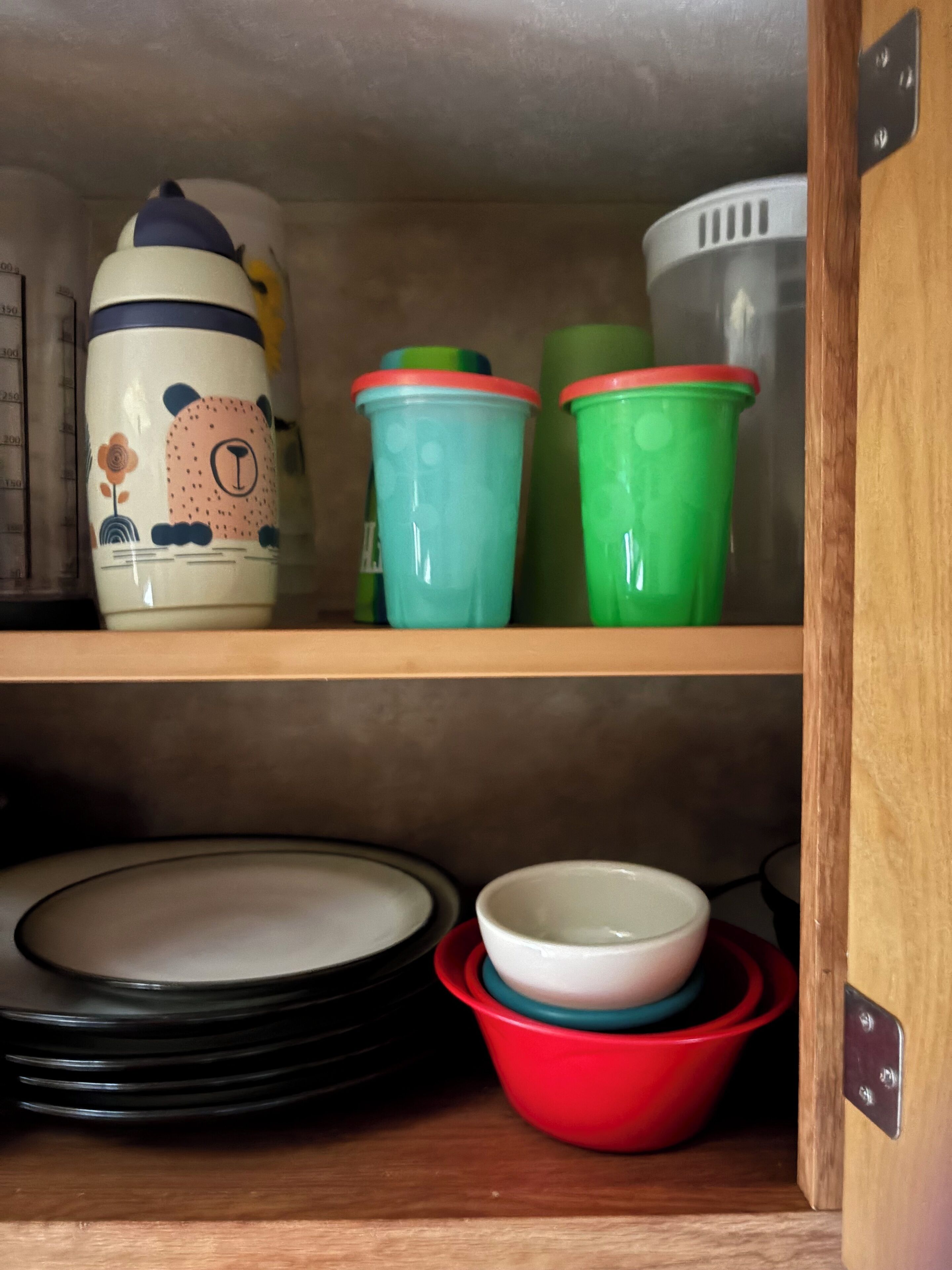 Una repisa de cocina con diversos utensilios, incluyendo una taza de cerámica con diseño floral, vasos de plástico verde y rojo, y platos negros apilados.