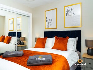 Camera d'albergo con un letto grande, biancheria arancione e bianca, una testiera blu scuro e due quadri con citazioni motivazionali appesi al muro.
