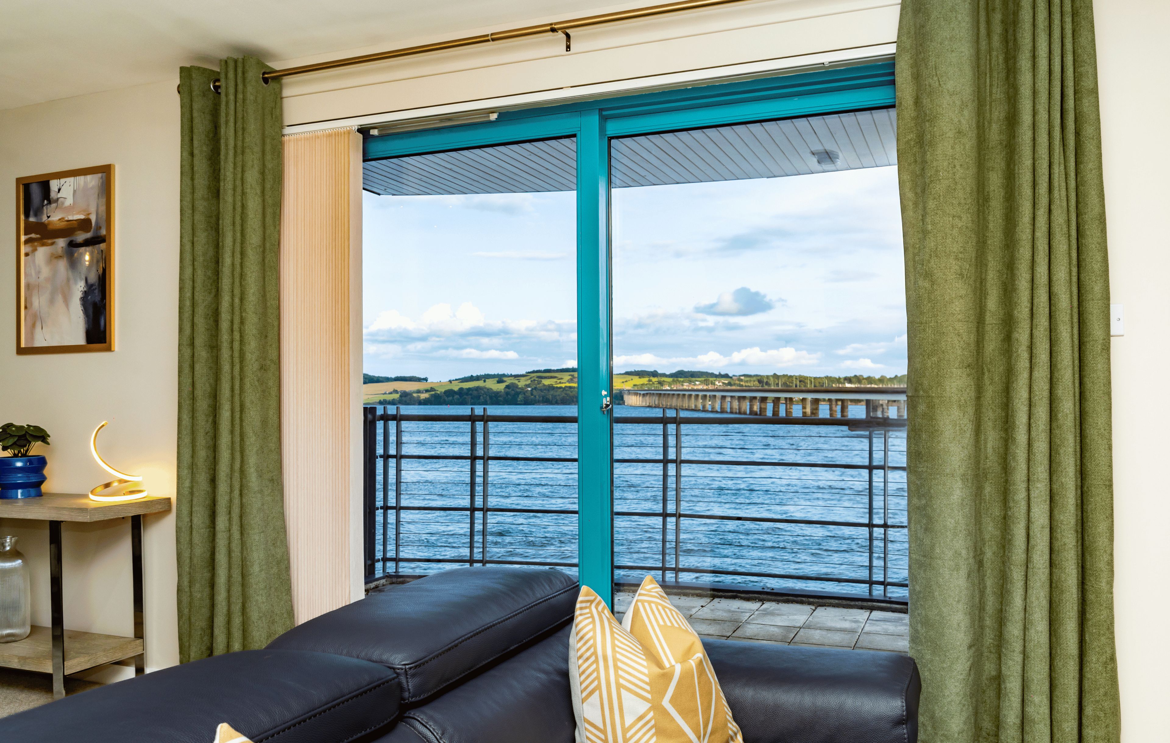 Una sala de estar con sofá de cuero negro, cortinas verdes y vista a una masa de agua con un muelle.