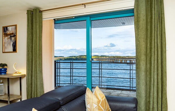Una sala de estar con sofá de cuero negro, cortinas verdes y vista a una masa de agua con un muelle.