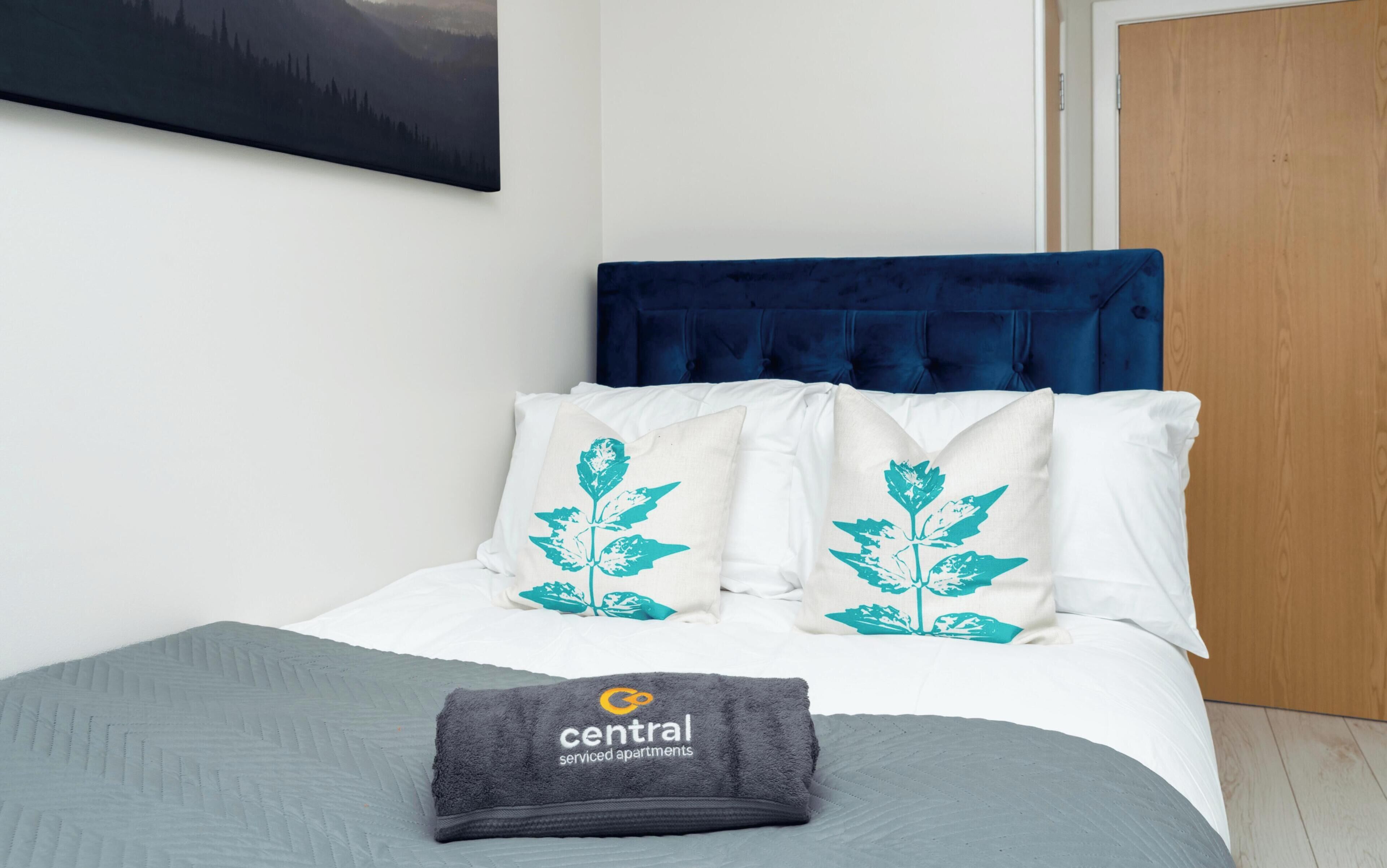 A bedroom with a bed, a blue headboard, white pillows with a plant design, a grey blanket, and a towel with a logo.