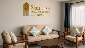 Un salon comprenant un canapé, deux fauteuils et une table basse. Un panneau est fixé au mur au-dessus du canapé, sur lequel on peut lire : « SRISUKSAN KLONG MUANG BEACH ».
