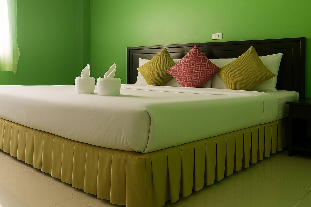 Un lit bien fait, recouvert d’une couvre-lit blanche et jaune, avec deux oreillers blancs et deux oreillers à motifs, dans une pièce aux murs et au sol verts.
