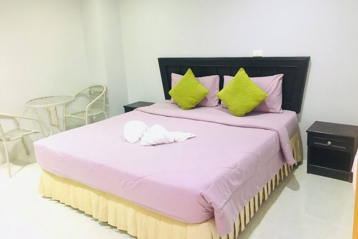 Un lit bien fait, avec une couvre-lit rose et deux coussins verts, une petite table ronde et une chaise, dans une pièce propre et blanche.