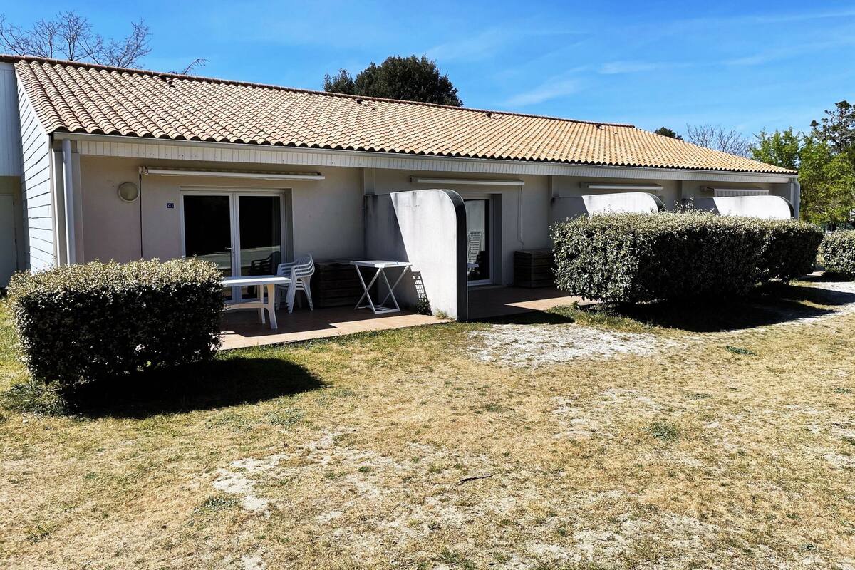 Un bâtiment d’un seul étage, doté d’un toit en tuiles, d’un patio avec une table et des chaises, et de deux arbustes bien entretenus.