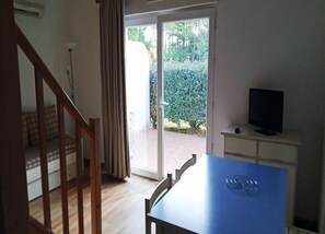 A room with a wooden staircase, a dining table with a blue tablecloth, a television on a cabinet, and a sliding glass door leading to a garden. - Apartment on Ile D'oleron Near the Beach (Saint-Georges-d'Oleron)