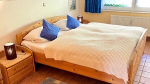 A wooden bed with a white blanket, blue pillows, a bedside lamp, a wooden nightstand, and a window with blue curtains. - Apartment in Lennestadt for 2 Person (Lennestadt)