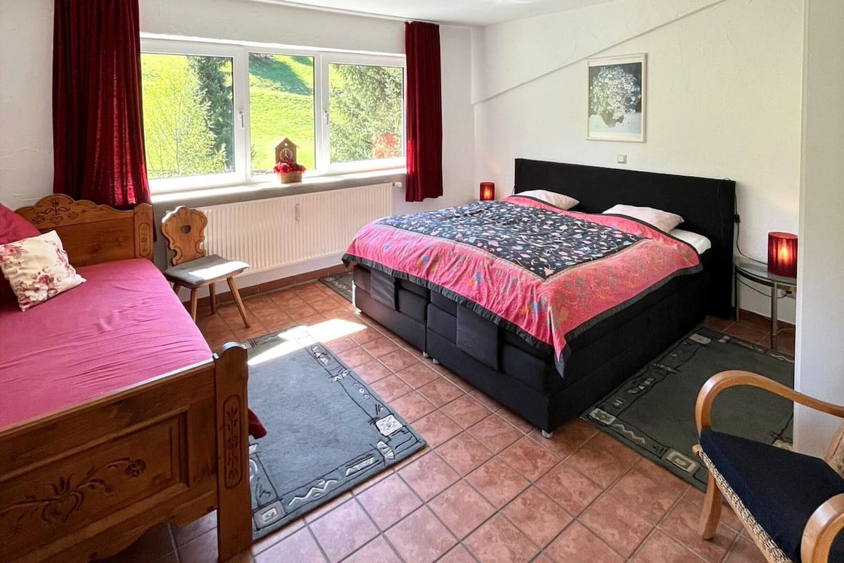 A bedroom with a bed, a chair, a window with curtains, a radiator, and a picture on the wall.