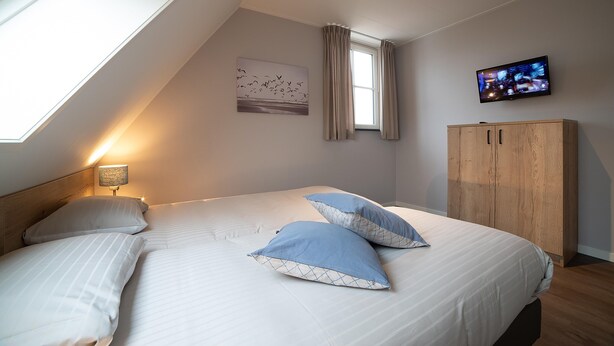 Een slaapkamer met een bed, kussens, een televisie, een raam en een houten garderobe.
