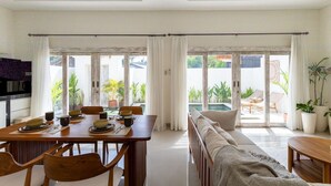 A modern dining area with a wooden table and chairs, a sofa, and a view of a pool and greenery. - Alinko Villa - 2BR Villa in Seminyak (Seminyak)