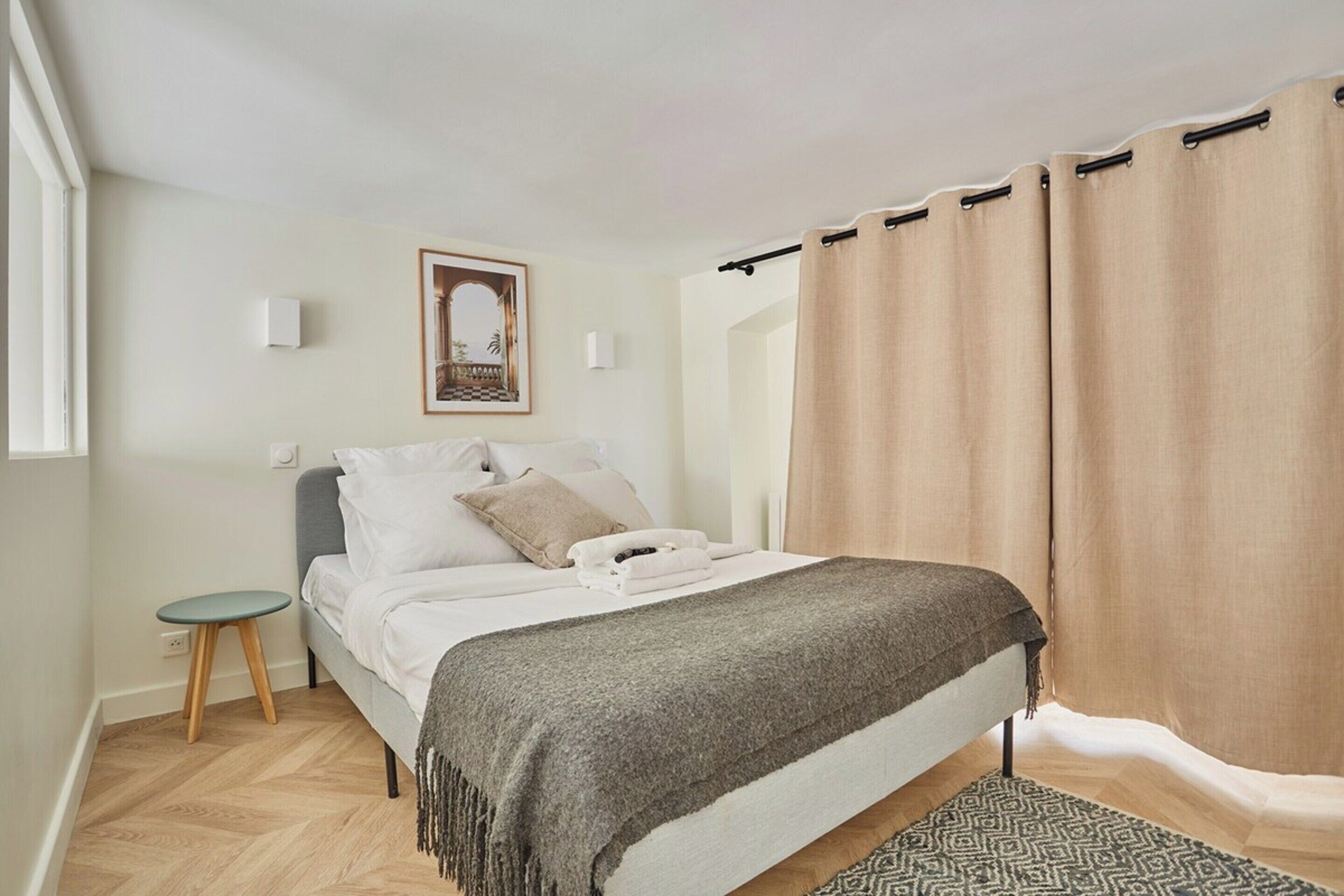 A bedroom with a bed, a small stool, a framed picture on the wall, and a window with curtains.