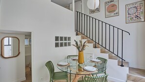 Un coin repas avec une table en verre, des chaises vertes et un vase contenant des plantes séchées. On aperçoit un escalier avec une rampe noire à l’arrière-plan.