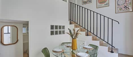 Un comedor con una mesa de vidrio, sillas verdes y un jarrón con plantas secas. Se observa una escalera con barandilla negra al fondo.