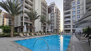 A swimming pool surrounded by lounge chairs and palm trees, with modern apartment buildings in the background. - Manzil - Cozy 1BR Waterfront Apartment  (Dubai)