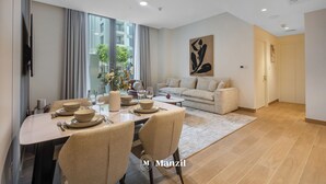 A modern dining area with a table set for two, a sofa, and a large window with curtains. - Manzil - Cozy 1BR Waterfront Apartment  (Dubai)