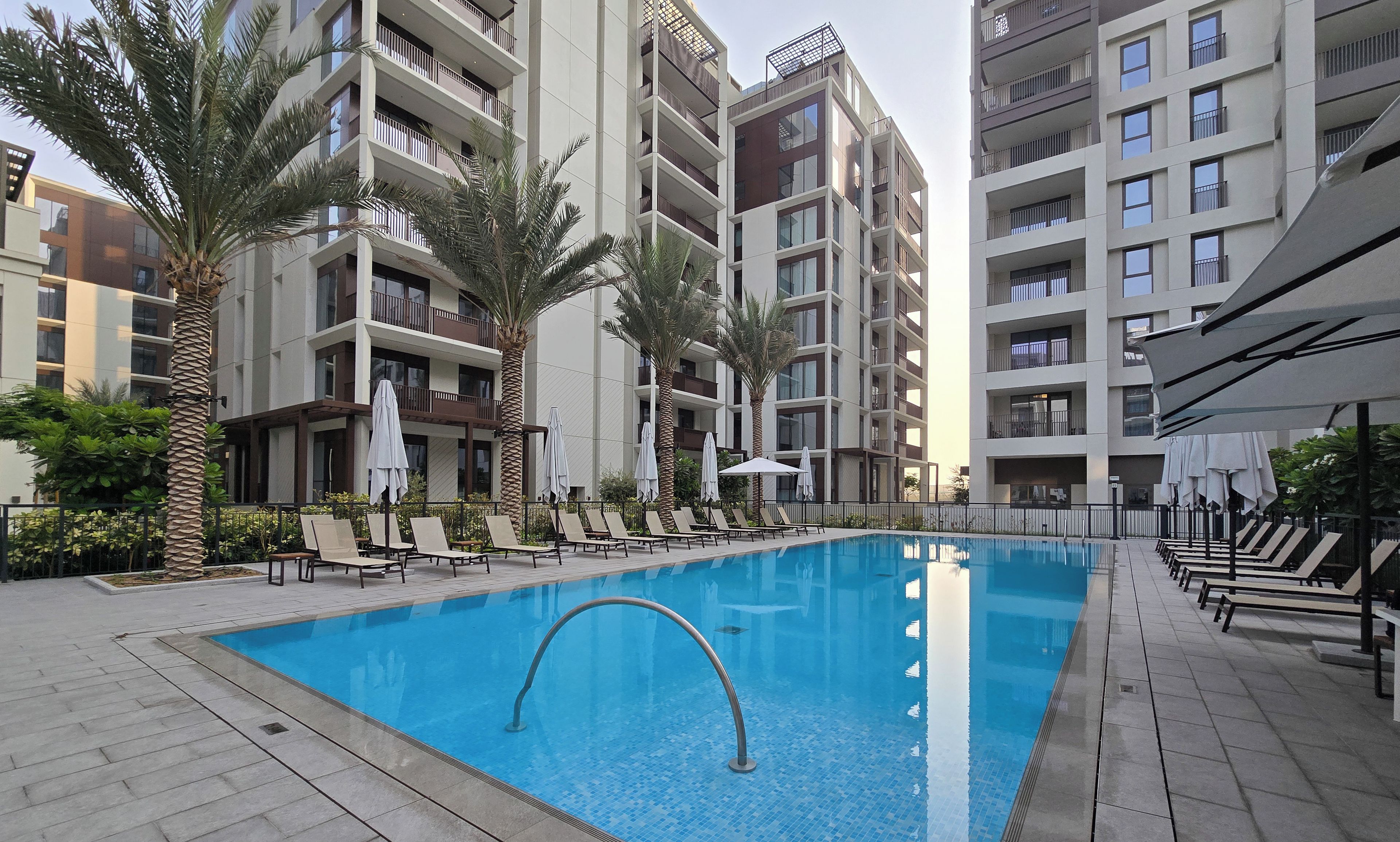 Una piscina rodeada de sillones de descanso y palmeras, con modernos edificios de apartamentos al fondo.