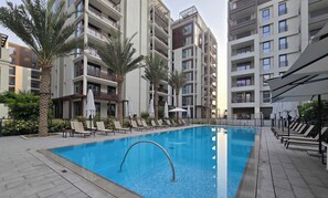 Een zwembad omringd door ligbedden en palmbomen, met moderne appartementen op de achtergrond.