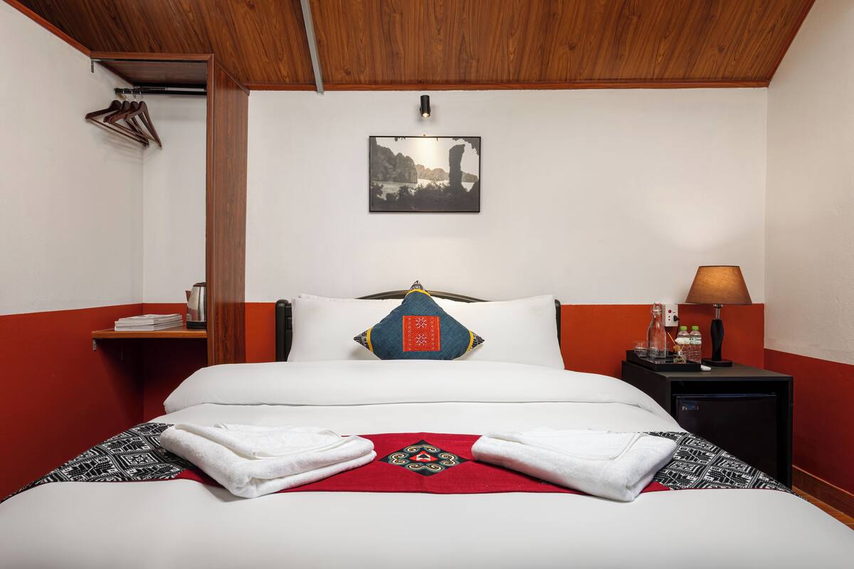 Un letto rifatto con una coperta a letto a motivi rossi e neri, cuscini bianchi e un cuscino blu con un disegno geometrico. Si nota un comodino con una lampada, un quadro appeso alla parete e un attaccapanni con grucce.