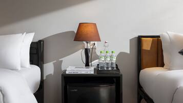 Ein Hotelzimmer mit zwei weißen Betten, einem Nachttisch mit Lampe, einem kleinen Kühlschrank und einem Tablett mit Wasserflaschen und einem Buch.
