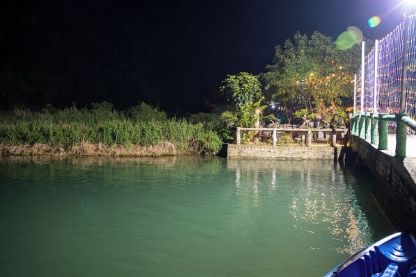 Escena nocturna de un río con un puente, vegetación e iluminación con luces colgantes.